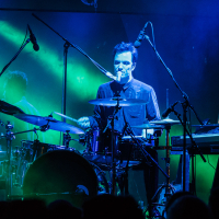 A drummer sits at a drum set on stage during a live performance at Piens Fest, illuminated by vibrant blue and green stage lights. Several microphones surround the setup, and a keyboard is to the drummer’s right with a laptop screen visible. The background includes faint outlines of other band members from 2014.