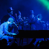 A musician plays a grand piano on stage under blue and green lights at Piens Fest 2014, while a brass ensemble in the background plays trombones and trumpets. The audience's silhouettes are visible in the foreground, and there is musical equipment to the left, suggesting an electric live performance.
