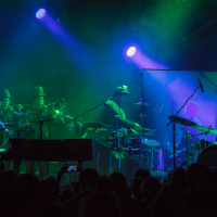 At Piens Fest 2014, a band performs on stage under blue and green lights. The scene shows a singer at a piano on the left, with musicians playing brass instruments and drums. The audience is visible in the foreground as silhouettes, and the stage has a vibrant, energetic atmosphere with spotlights illuminating the performers.