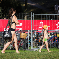 A woman in a black bikini walks barefoot on grass, carrying a black shoulder bag, accompanied by a young child wearing green shorts and a yellow hat. Behind them, bicycles are parked behind a wire fence, and a red Piens Fest 2014 sign with white graphics stands tall.