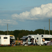 A group of people are enjoying a camping trip in a grassy field with two white RVs and a tent. They are sitting and standing around, engaging in conversation and activities reminiscent of Positivus AB 2008. The sky is partly cloudy, and there are trees and utility poles in the background.