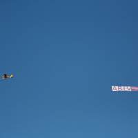 A small yellow airplane is flying in a clear blue sky, towing a long rectangular banner. The banner is partially readable, displaying the letters "ABLY" and some additional text that is not fully legible, possibly promoting Positivus 2008. The scene is set against a plain backdrop with no clouds visible.