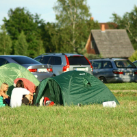 Two green camping tents are set up on a grassy field. Two people sit on the grass near the tents, with one holding a red and white checkered cloth, reminiscent of a Positivus festival scene. Multiple parked cars are in the background, along with trees and houses under a clear sky.