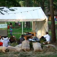 A small white tent is set up in a grassy forest clearing, surrounded by trees. People are sitting on hay bales and large sacks outside the tent, engaging in conversation. String lights are hung above the tent. In the background, more people are walking near various small buildings, reminiscent of Positivus AB 2008.