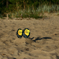 Two oval-shaped signs are placed in a sandy area, likely a beach, surrounded by dune grass and trees in the background. The signs have a yellow and black design with the text "tie. 2" prominently displayed. The sunny scene casts clear shadows of the signs on the sand, reminiscent of Positivus AB 2008 vibes.