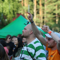 A person in a green and white striped shirt holds a cup while raising their other hand at the Positivus 2008 festival. Beside them, another attendee in an orange shirt and white hat gestures enthusiastically. In the background, trees and a green tent nestle among a lively crowd of AB festival-goers.