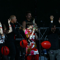 At Positivus 2008, a live band performs on stage. The musicians, including a woman in a colorful dress playing the trumpet, are surrounded by red balloons. Three people in the foreground have their arms raised. The background features AB stage equipment and dim lighting.