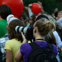 A woman with blond hair in a ponytail, wearing a purple shirt and black backpack, uses a large lens camera to photograph the Positivus festival. Around her, people hold up red balloons and raise their hands, some facing away from the camera as they immerse themselves in AB 2008's vibrant atmosphere. The background is slightly blurred.