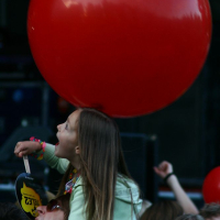 At Positivus, a young child with long hair, wearing a light green sweater, sits on an adult's shoulders in a crowd. The child looks up excitedly at a large red balloon above their head. They hold a small, round yellow sign. The background is dark, with blurred elements suggesting the lively atmosphere of AB 2008.