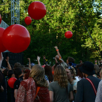 A crowd of people at the Positivus 2008 outdoor concert is enjoying the event. Large red balloons are being tossed among the attendees. The background is filled with lush green trees, and people are dressed in casual clothing. The atmosphere appears lively and festive.