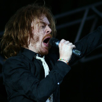 A singer with long, tousled hair passionately performs on stage at Positivus 2008, holding a microphone close to his mouth. He is wearing a black shirt and a light-colored tie. His right arm is raised, and his face is animated with emotion. The background is dark with part of a metal structure visible.