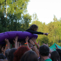 A crowd of people, many with raised hands, holds up a person riding a purple inflatable raft. The individual on the raft points outward with their right hand. The outdoor setting, surrounded by trees and vibrant foliage, hints at the lively atmosphere of an event like Positivus or AB 2008.