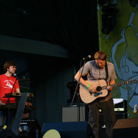 Two musicians perform on an outdoor stage at AB 2008 Positivus. The musician on the right plays an acoustic guitar and sings into a microphone, while the musician on the left, in a red shirt with glasses, plays a keyboard. Equipment and speakers surround them against a backdrop of bright, abstract design.