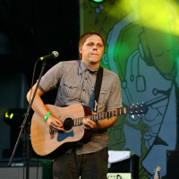 A person stands on an outdoor stage at AB 2008, playing an acoustic guitar. They are wearing a checked shirt and cargo pants. Behind them, a yellow and green abstract backdrop is illuminated by stage lights. A microphone stand is positioned in front of the person, and audio equipment is visible in the background.