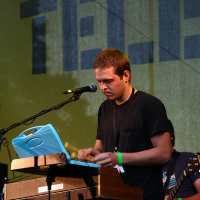 A man standing and playing a keyboard on a wooden stand under a microphone at Positivus. Wearing a black shirt and a wristband on his left hand, he captivates the crowd. In the background, an obscured band member plays guitar, with "TELE" visible on the 2008 backdrop behind them.