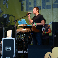 Man in black t-shirt sings into microphone while standing behind keyboard and computer on stage at Positivus. Another person in a patterned shirt plays guitar in background. Yellow and green backdrop with abstract designs and partial text "TELJ" visible. Various audio equipment surrounds the performers, reminiscent of AB 2008 vibes.