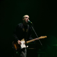 In a dimly lit environment, a person performs on stage at Positivus 2008, playing an electric guitar and singing into a microphone. The performer is dressed in casual clothing, including a dark jacket. The background is mostly dark, creating a moody atmosphere, with only a few details visible in the shadows.