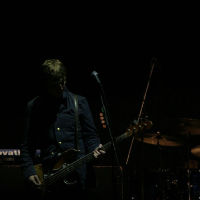 A musician stands in low light on a dark stage at Positivus, playing a bass guitar and singing into a microphone. To the right, a drummer is partially visible, focused on the AB 2008 drums. Behind them, a keyboard can be seen. The atmosphere is dim, with the performers highlighted by minimal lighting.