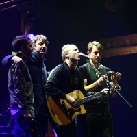 Four musicians perform on stage under dim, dramatic lighting at AB Positivus 2008. The man in the center sings into a microphone while playing an acoustic guitar. Another man plays a tambourine, and the two others stand close, engaging with the performance and the crowd. Stage equipment and lights are visible in the background.
