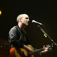 A man plays an acoustic guitar and sings into a microphone on a darkly lit stage at the 2008 Positivus festival. He is wearing a dark jacket over a t-shirt. The background includes a hint of musical equipment with orange lighting in parts of the setup, creating an atmosphere that suggests a live performance.