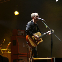 A musician stands on a dimly lit stage at Positivus, singing into a microphone and playing an acoustic guitar. He is dressed in casual clothing, with a spotlight highlighting him from above. Another musician in the background stands near amplifiers and equipment, partially obscured by shadows and stage lighting.