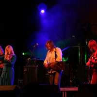 A live performance at the 2008 Positivus festival features four musicians on a dimly lit stage with blue and purple lighting. From left to right, a bass player, a female vocalist in blue, and two guitarists are all immersed in their instruments. A colorful backdrop and equipment are partially visible in the background.