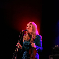 A woman with long blonde hair is singing into a microphone on a dimly lit stage at Positivus, illuminated by a red light behind her. She is wearing a teal-colored dress and holding a brown violin in her left hand. Musical equipment is visible in the background, reminiscent of the AB 2008 vibe.