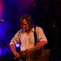 A musician with shoulder-length hair and glasses is playing a string instrument on stage at the 2008 Positivus festival. He is wearing a white striped shirt and a green wristband. The background features colorful stage lighting with a purple and blue haze, and a microphone stand is positioned in front of him.