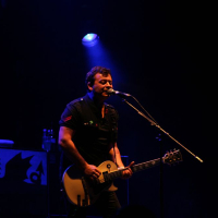 A musician stands on stage under dim blue and purple lighting at Positivus AB 2008, playing an electric guitar and singing into a microphone. He wears a dark shirt and looks focused on his performance. In the background, there is a black case with white designs on it. The overall atmosphere is moody and concert-like.