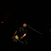 A guitarist performs passionately on stage at Positivus 2008, holding a white electric guitar and leaning into a microphone. The stage is dimly lit with a few orange and red spotlights, creating a moody atmosphere. The dark background emphasizes the musician as the focal point.