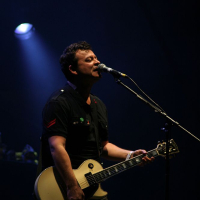 A man performing on stage at Positivus plays an ivory-colored electric guitar and sings into a microphone. Illuminated by a single spotlight against a dark background, he wears a black shirt with red and green patches. His expression is focused, and various equipment is visible in the dim background.