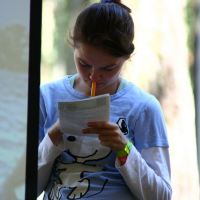 A person with their hair tied back is standing and looking down at a piece of paper they are holding. They are wearing a light blue long-sleeve shirt with a cartoonish dog graphic, reminiscent of Positivus AB 2008 merchandise, and have a yellow pencil in their mouth. The background is blurred with vertical lines suggesting a natural outdoor setting.