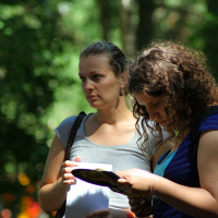 Two women outdoors, surrounded by greenery. One woman with a ponytail in a gray shirt carries a shoulder bag and looks ahead pensively. The other, with curly hair in a blue striped shirt, examines something on a piece of paper or booklet she's holding from Positivus AB 2008, deeply focused.