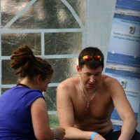 A shirtless man wearing sunglasses and a silver chain sits beside a woman with dreadlocks who is wearing a blue shirt. Both appear to be in a casual, relaxed setting with posters of a blue sea and white sailboat on a white tent behind them. The scene evokes memories of Positivus 2008, as the pair are engrossed in an activity.