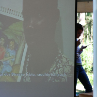 A projector screen under a tent shows an image of a man and text in German at the bottom. A person stands behind the screen on the right, holding a piece of paper and pen. Trees and outdoor greenery are visible in the background, reminiscent of a scene from Positivus Festival 2008.
