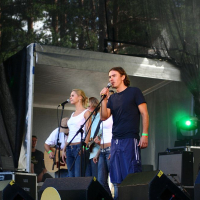 A man and a woman are singing into microphones on an outdoor stage at Positivus, with multiple performers and equipment behind them. The man is wearing a dark T-shirt and shorts, while the woman sports a white top and jeans. Green stage lights illuminate the area, with trees providing a scenic backdrop.