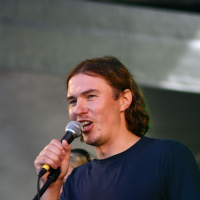 A person with shoulder-length brown hair stands in front of a microphone. They are wearing a plain dark blue T-shirt. The background is blurred, focusing attention on the individual who appears to be speaking or singing, reminiscent of performances at Positivus 2008.