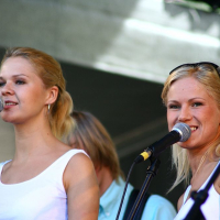 Two young women with blonde hair are singing into microphones on a stage at Positivus 2008. They are wearing white tops and positioned in front of a blurred background that includes another individual partially visible between them. Both women have smiling expressions and appear to be enjoying the performance.