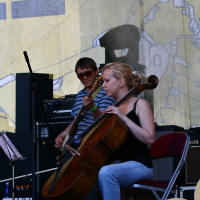 A person with a cello is seated in mid-performance, accompanied by another playing an electric bass guitar in the background. They are on a stage at Positivus, surrounded by various musical equipment and a large illustrated backdrop. The cellist appears focused, and the guitarist stands behind them.