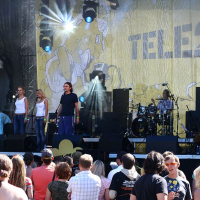 A band performs on an outdoor stage with a yellow backdrop displaying the "Tele2" logo and abstract designs. At Positivus, four vocalists stand in front, next to other musicians, including a drummer and cellist. The audience, dressed in casual clothing, watches the performance under sunny skies.
