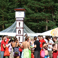 At the Positivus festival in 2008, a group of people, mostly in swimwear and summer clothes, gather around a tall, white cuboidal signpost featuring the brand "Laima" with various chocolate product adverts. A white tent and towering pine trees provide the backdrop. Some individuals hold red bags, while a few wear towels.