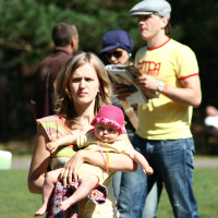 A woman with blond hair carries a baby wearing a pink hat and holding a patterned cloth in a sunlit park. Behind them, a man in glasses and a hat reads an AB 2008 newspaper next to a woman in a blue outfit and sunglasses. Other individuals walk in the background, with Positivus energy among the trees and grass.