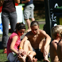 A shirtless man and a girl in a pink shirt sit on the grass at Positivus 2008; the girl places her hand on his ankle. Both sport yellow wristbands. In the background, another person in a brown shirt and gray pants is partially visible, with additional people blurred further back.