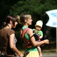 Two women are standing outdoors in a grassy area at Positivus. One woman is holding a baby wrapped in a colorful sling. Both women are holding frisbees and looking in the same direction. The baby, wearing a yellow beanie and green outfit, gazes curiously as trees and an AB 2008 sign stand in the background.