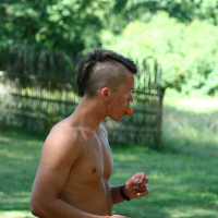 A shirtless person with a mohawk hairstyle stands outside in a grassy area, holding a small object in one hand while biting into an orange item, possibly a fruit. Wearing a wristband and shorts with a bracelet on the right wrist, they exude vibes straight out of Positivus. A wooden fence and trees are visible in the background.