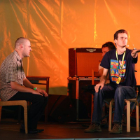Two men are on a stage with an orange backdrop at the 2008 Positivus AB festival. One is sitting on the left in a plaid shirt, facing the other man on the right. The man on the right is seated, wearing a black graphic t-shirt, and extending his right arm forward, slightly pointing. Musical equipment is visible in the background.