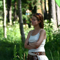 A woman with shoulder-length brown hair stands in a forested area at Positivus 2008, looking to the side with arms crossed. She wears a light-colored tank top and patterned brown pants, with a beige bag slung across her hips. Sunlight filters through the trees, creating a dappled light effect on the greenery.