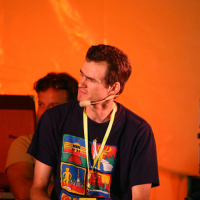 A man wearing a navy blue T-shirt with colorful graphics and a yellow lanyard around his neck sits in front of an orange background during the 2008 Positivus festival. He has a microphone headset and appears to be listening intently. Another person is partially visible in the background next to musical instruments.