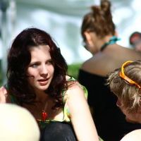 A young woman with long, wavy dark hair is sitting on the grass in a lively outdoor setting, talking to a young man wearing sunglasses on his head. The atmosphere is as relaxed and sunny as Positivus 2008, with other people casually sitting and engaging in conversation in the background.