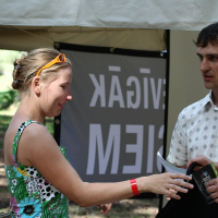 A woman in a floral dress and sunglasses on her head is smiling and interacting with a man in a white, patterned shirt under a tent labeled "Positivus." The man is holding documents, while the woman, sporting a red wristband on her left hand, looks cheerful. Perhaps they’re reminiscing about AB 2008.